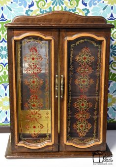 an ornate wooden cabinet with glass doors and gold trimmings on the door, in front of a floral wallpaper