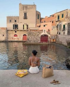 a woman sitting on the edge of a body of water next to buildings and a book