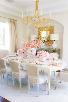 a dining room table set with pink flowers and place settings in front of a chandelier