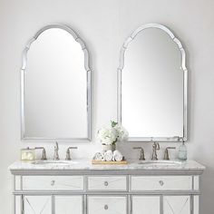 two mirrors are on the wall above a double sink vanity with white flowers in front of it