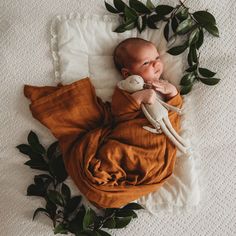 a baby wrapped in an orange blanket laying on top of a white bed next to green leaves