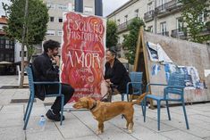 two people sitting on chairs with a dog in front of them and a large poster behind them