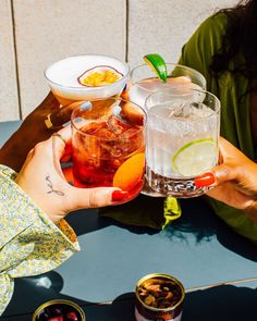 two people holding up glasses filled with different types of drinks and fruit on the table