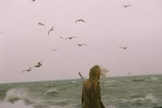 a woman is walking on the beach with birds flying above her by an ocean shore