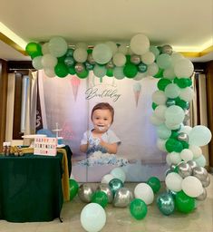 a baby is sitting in front of a birthday cake and balloon arch with green, white and silver balloons