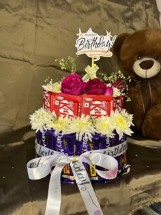 a teddy bear sitting next to a birthday cake with flowers and chocolates on it