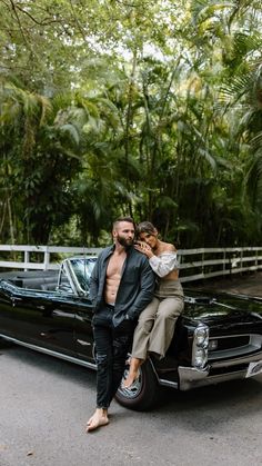 a man and woman sitting on the hood of a classic car in front of trees