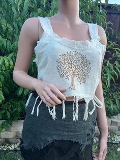 Introducing our Tree Of Life Tassel Top. This beautifully crafted crop top features a traditional Tree of Life motif, meticulously hand block printed on soft, breathable cotton. The intricate design is complemented by playful tassels, adding a touch of bohemian charm. The top has unstitched edges to make it look more natural and rugged. Perfect for warm weather, this top pairs effortlessly with high-waisted jeans, skirts, or shorts for a relaxed yet stylish look. Whether you're heading to a fest Embroidered Cropped Tops For Vacation, Beach Embroidered Cotton Crop Top, White Cotton Top With Back Tassel Tie-up, Summer Crop Top With Frayed Hem, Cropped Cotton Festival Top, Summer Cropped Top With Frayed Hem, Cropped Cotton Top For Festival, Summer Cropped Crop Top With Frayed Hem, Bohemian Cotton Tops With Frayed Hem