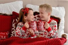 two children in matching christmas pajamas holding red mugs and smiling at each other on a bed