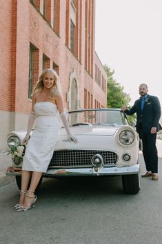 a man and woman standing next to an old fashioned car in front of a brick building