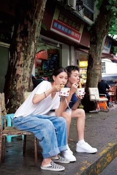 two people sitting on a bench eating food and drinking coffee in front of a store