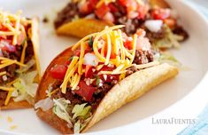 three tacos on a white plate topped with shredded cheese and toppings, sitting on a blue table cloth