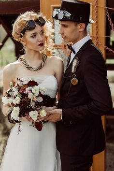 a man and woman standing next to each other in front of a wooden structure holding flowers