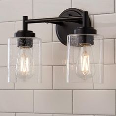 two light fixtures with clear glass shades on the wall in a white tiled bathroom area