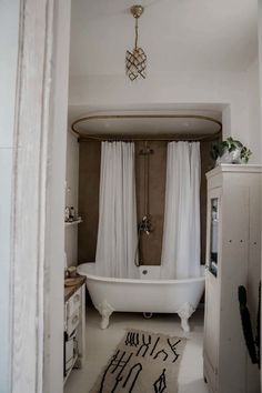 a bath tub sitting under a window next to a white curtained bathroom sink and toilet