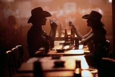 two people sitting at a table with beer bottles in their hands and one person wearing a cowboy hat