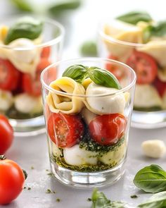 three small glasses filled with pasta and tomatoes on top of a table next to basil leaves