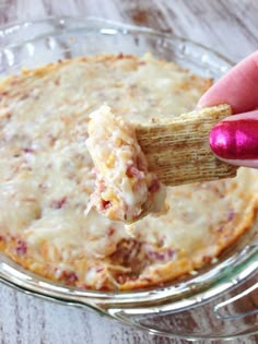 a hand holding a cracker over a casserole dish with cheese and meat