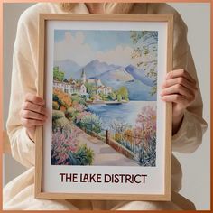 a woman holding up a framed painting with the words the lake district in it's center