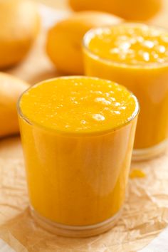 two glasses filled with orange juice sitting on top of a table next to mangoes