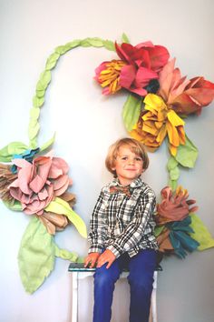 a young boy sitting on a stool in front of a paper flower wall art piece