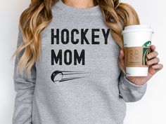 a woman holding a cup of coffee and wearing a hockey mom sweatshirt