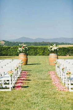 an outdoor ceremony setup with white chairs and flower petals on the aisle, surrounded by wooden barrels