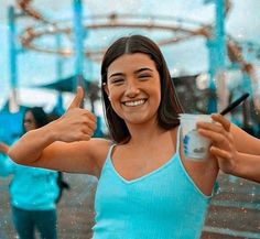 a woman in a blue tank top holding a drink and giving the thumbs up sign