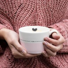 a woman holding a cup with a heart on it in her hands and wearing a pink sweater