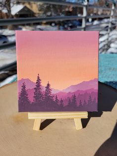 an easel with a painting on it sitting on top of a wooden table in front of snow covered mountains