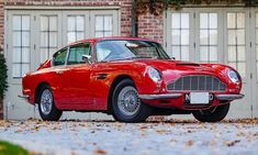 an old red sports car parked in front of a brick building with two garage doors