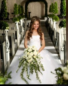 a woman in a white wedding dress holding a bouquet and standing next to rows of pews