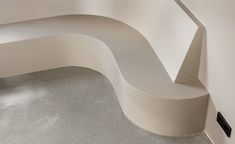 a curved white bench sitting on top of a cement floor next to a wall and window