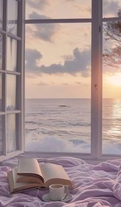 an open book sitting on top of a bed in front of a window overlooking the ocean