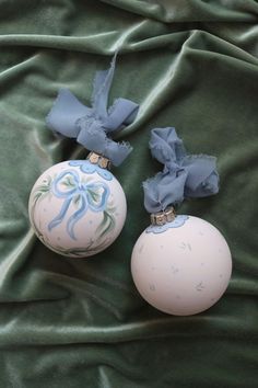 two ornaments with bows on them laying on a green cloth covered bed sheet, one is white and the other is blue