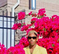 a man in a yellow suit and sunglasses standing next to pink flowers with a brick wall behind him