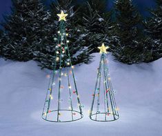 two metal christmas trees with lights in the middle of snow covered ground and evergreens behind them