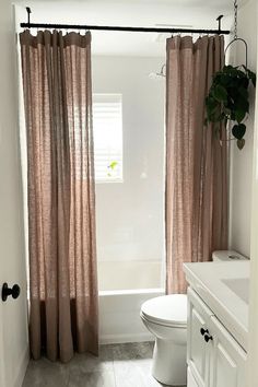 a white toilet sitting next to a bath tub in a bathroom under a window with pink curtains