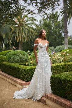 a woman in a white wedding dress standing on some steps near bushes and trees with palm trees behind her