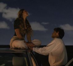 a woman sitting on the hood of a car talking to a man standing next to her