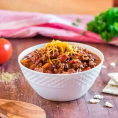 a white bowl filled with chili and cheese on top of a wooden table next to crackers