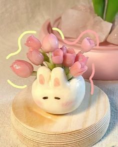 a small white vase with pink flowers in it on top of a wooden coaster next to a potted plant