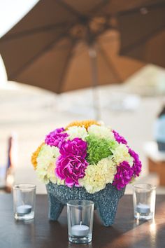 an arrangement of flowers is placed on a table with shot glasses and umbrella in the background