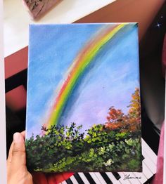 someone is holding up a painting with a rainbow in the sky and trees on it