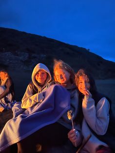 three women sitting next to each other in front of a campfire at night time