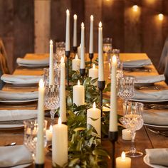 a long table with white candles and greenery on the top, surrounded by other place settings