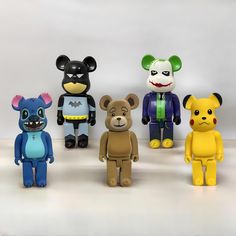 five different sized toy bears are lined up on a white surface, one is wearing a batman costume and the other has a clown mask