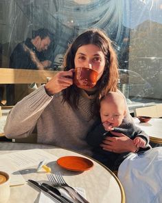 a woman sitting at a table holding a baby and drinking from a mug in front of her