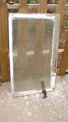 a broken glass window sitting on the ground next to a wooden fence with a screwdriver in it