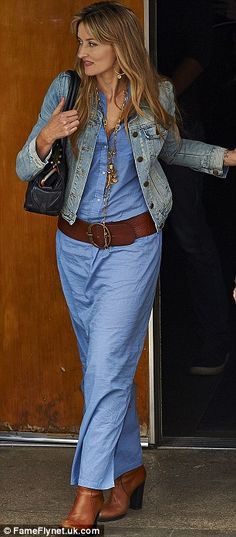 a woman in a blue dress and jean jacket is walking out of a building with her hand on her hip